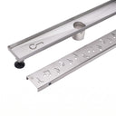 A stainless steel linear shower drain from megabai with a removable musical note grate and adjustable leveling legs.